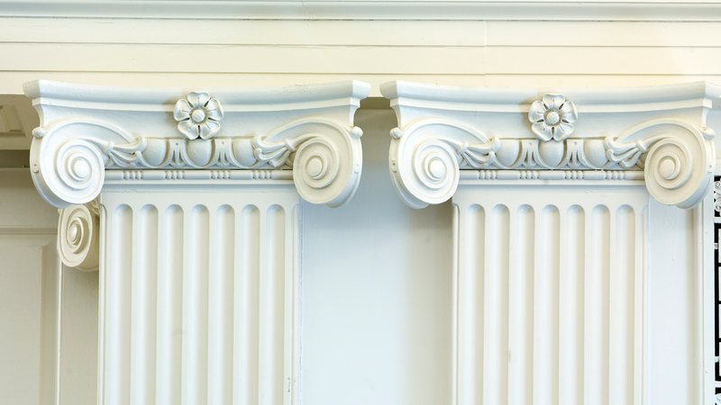 Two white Ionic pilasters with scrolled capitals and fluted shafts against a pale cream wall