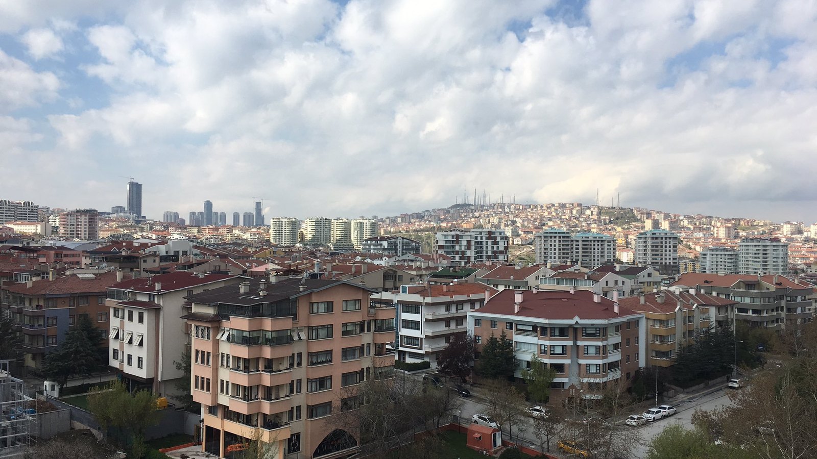 Red-roofed apartment blocks spreading towards distant high-rise towers and a hilltop under a cloudy sky in Ankara