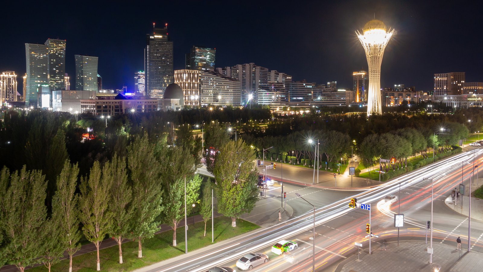 Astana at night with the illuminated Bayterek tower, lit high-rises and light trails on a wide avenue