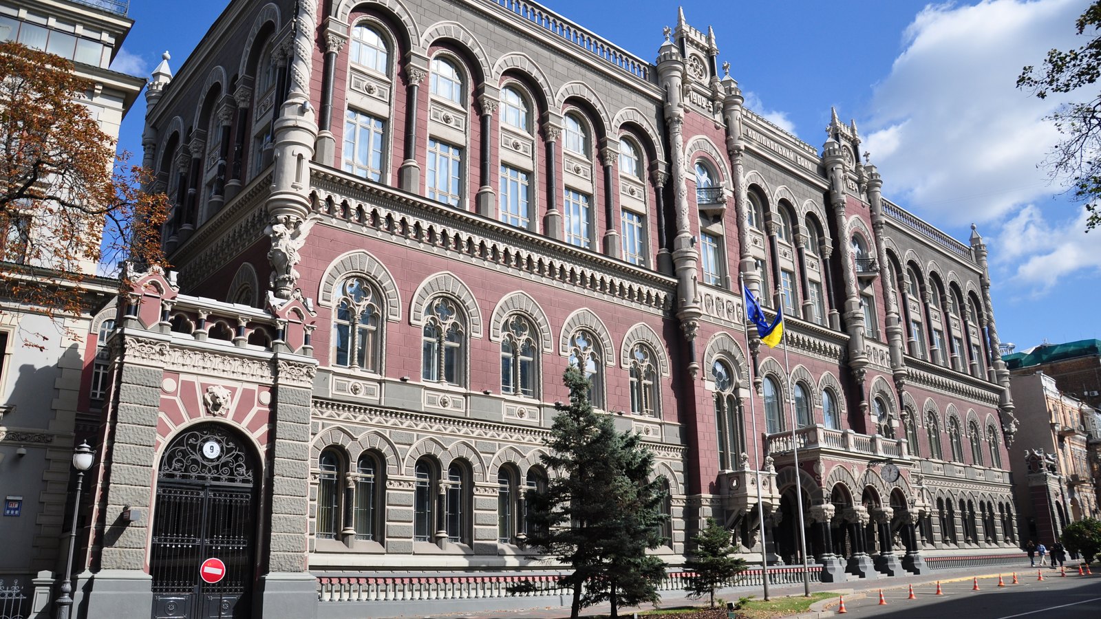 Ornate pink and grey bank building with arched windows, turrets and a blue and yellow flag under a cloudy blue sky