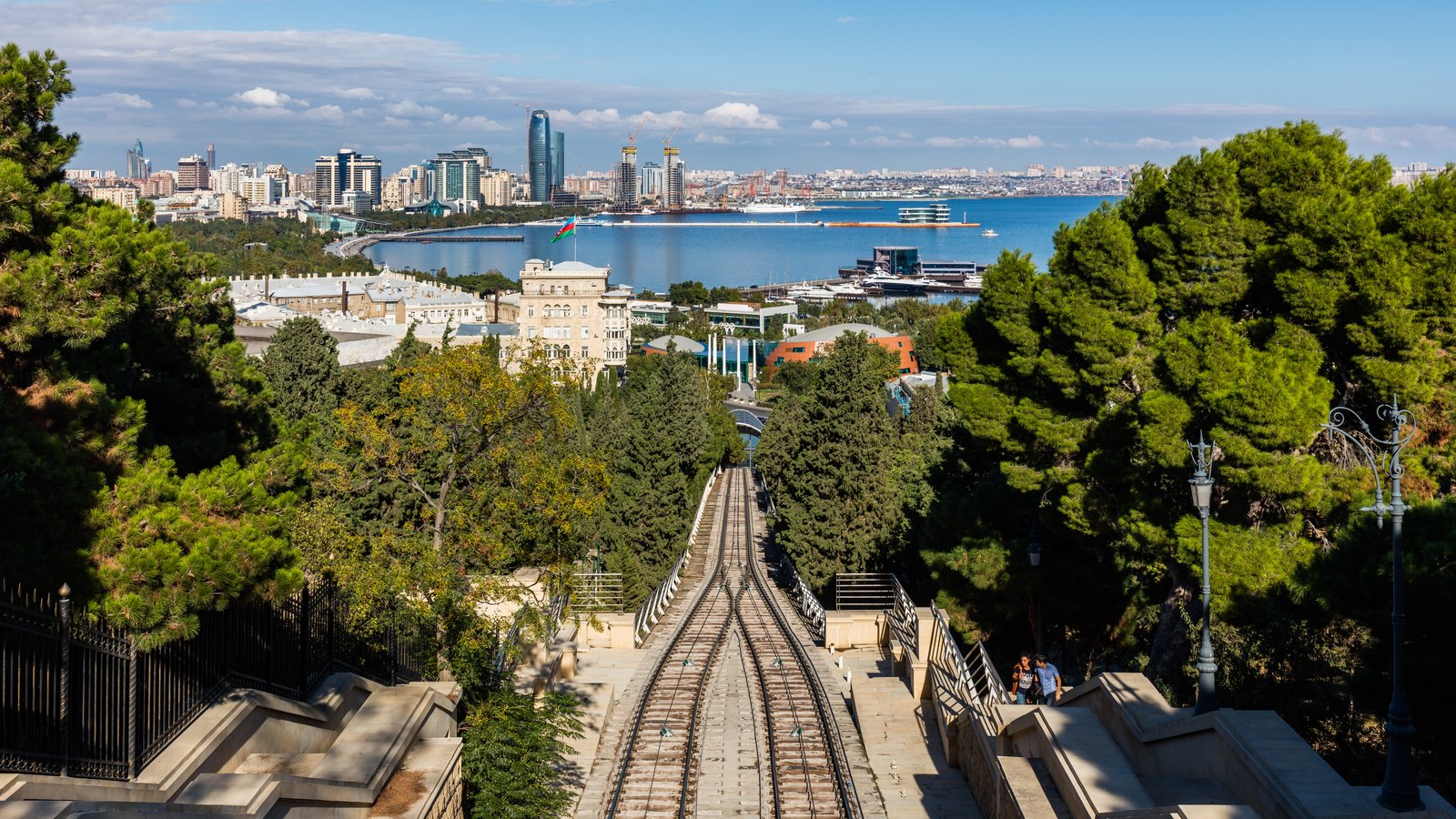 Funicular tracks run steeply downhill through trees towards a bay lined with high-rise towers