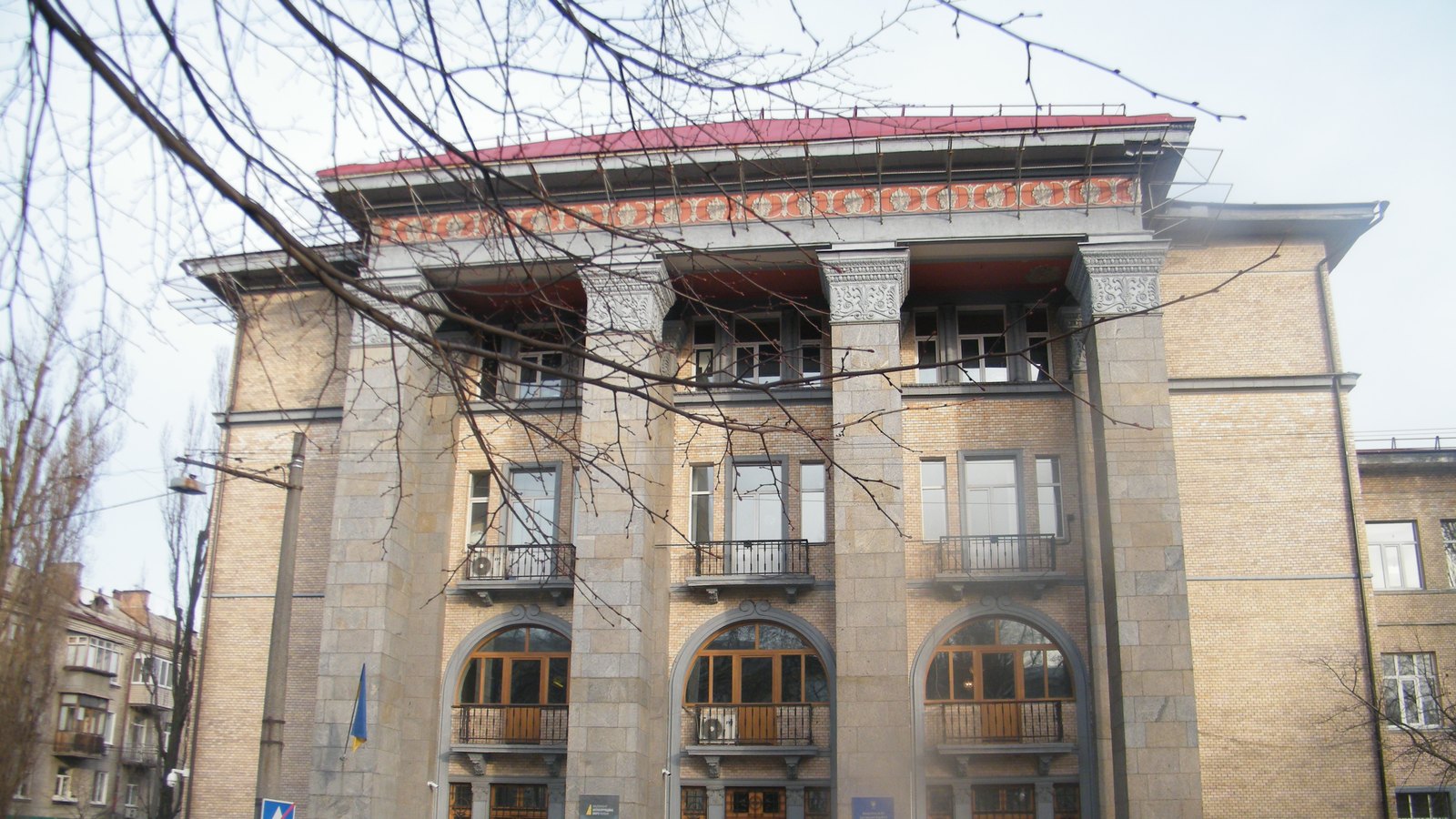 A stone-clad building with arched windows and balconies, a Ukrainian flag on the facade, seen through bare branches