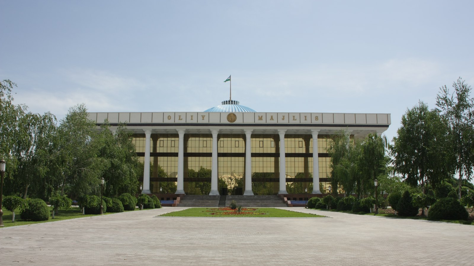 A white colonnaded building marked Oliy Majlis, with a small blue dome and a flag, faces a wide paved square