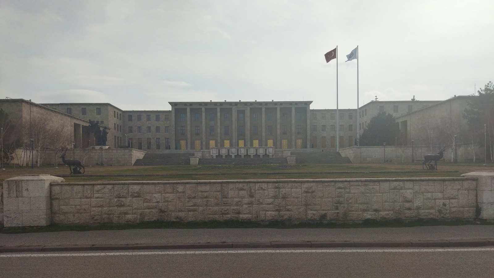 A long colonnaded stone parliament building behind a wide lawn and low wall, with two flags flying
