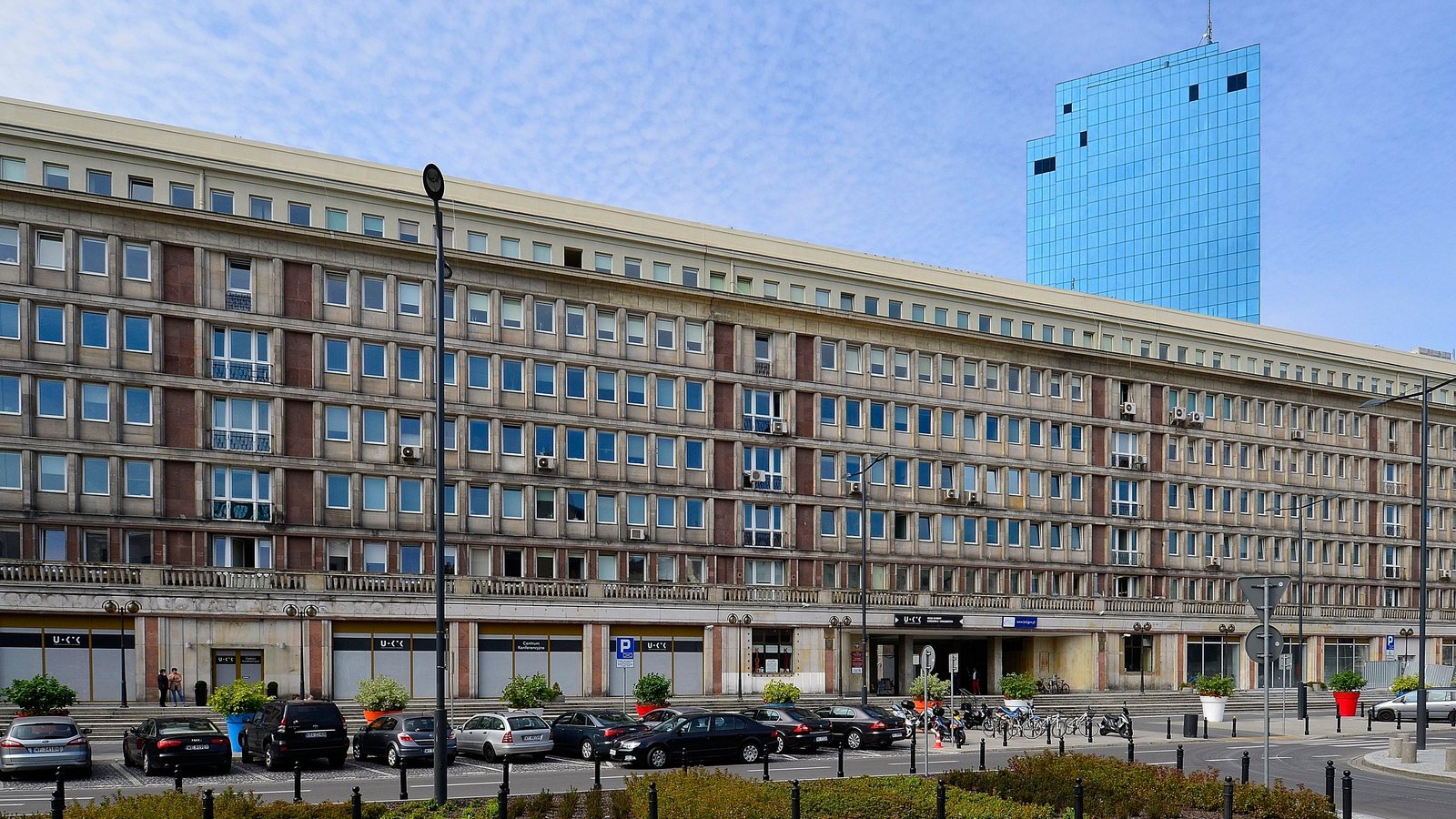 A long stone office block with cars parked in front and a blue glass tower rising behind it