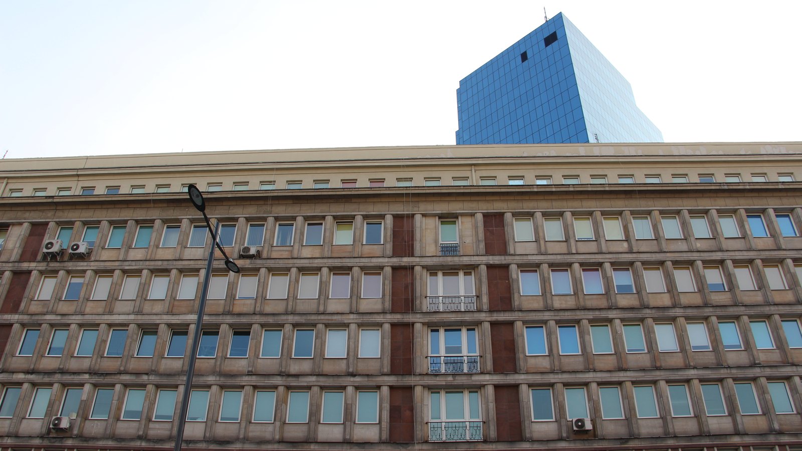 Upper floors of a stone office block with small balconies, with a blue glass tower rising behind