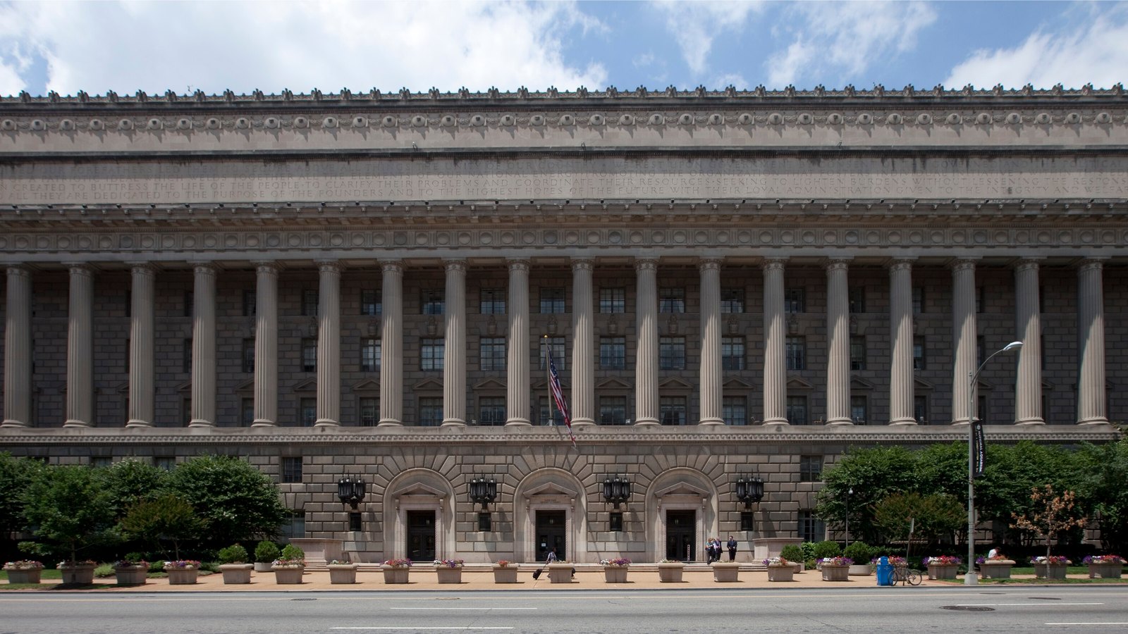 A long neoclassical stone building with a row of tall columns and a flag above the central entrance