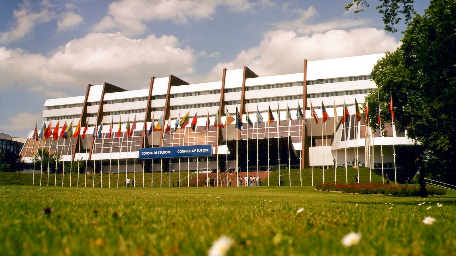 The long facade of the Palais de l'Europe behind a row of national flags and a lawn