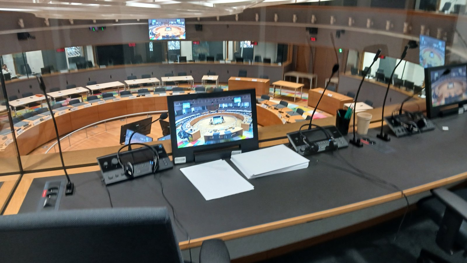 A view from an interpreting booth, with its microphones and screens, into a large meeting room with a ring of desks