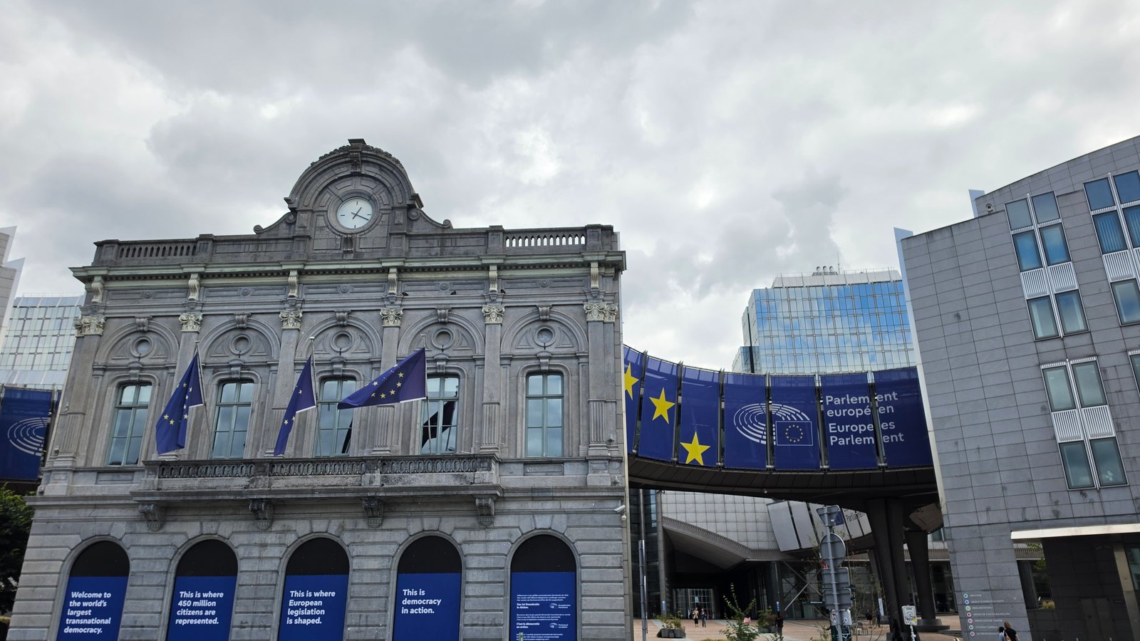 A historic station building with a clock and EU flags, between modern glass buildings of the European Parliament