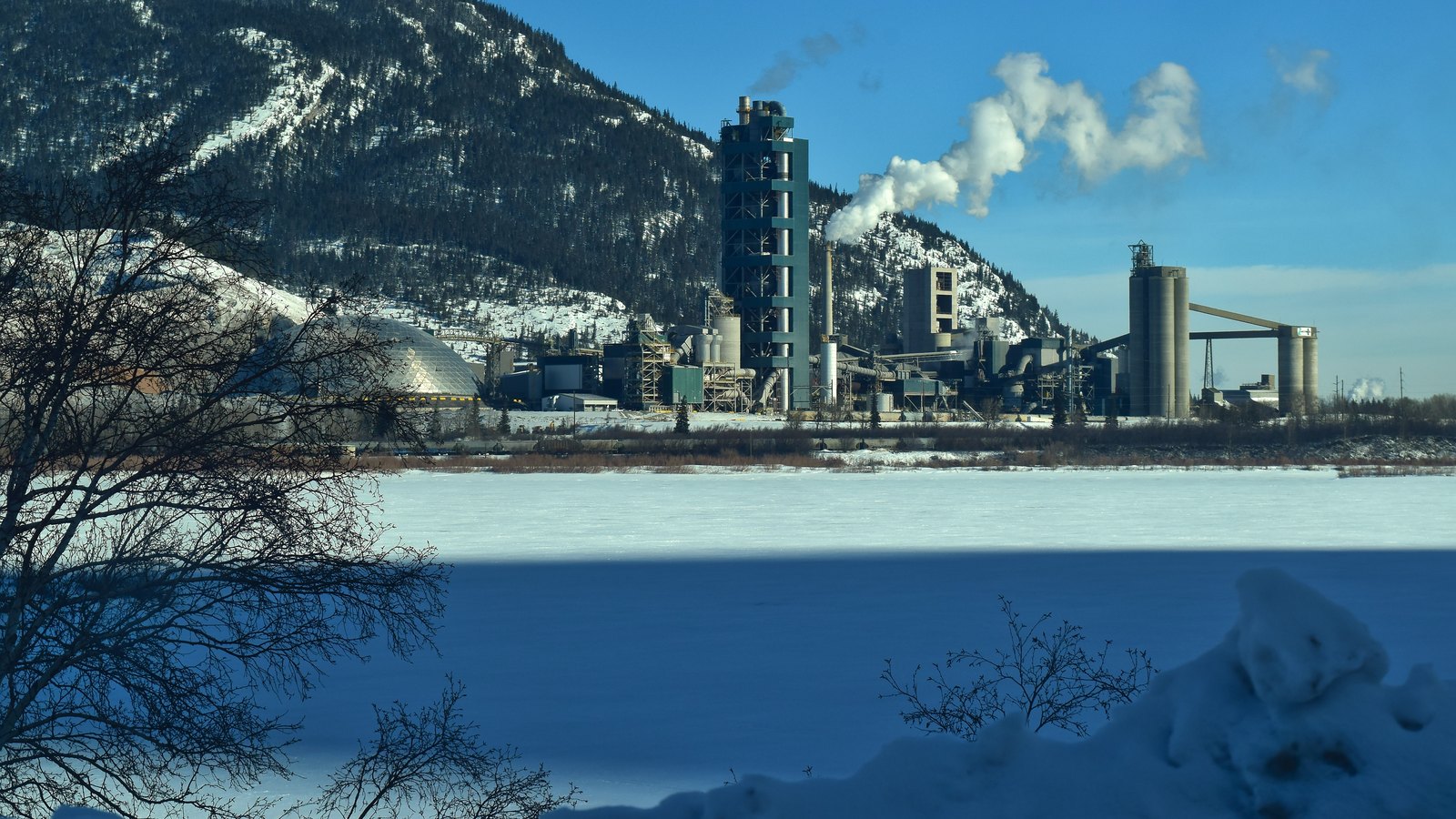 A cement plant releasing white steam at the foot of snowy mountains, beyond a frozen lake