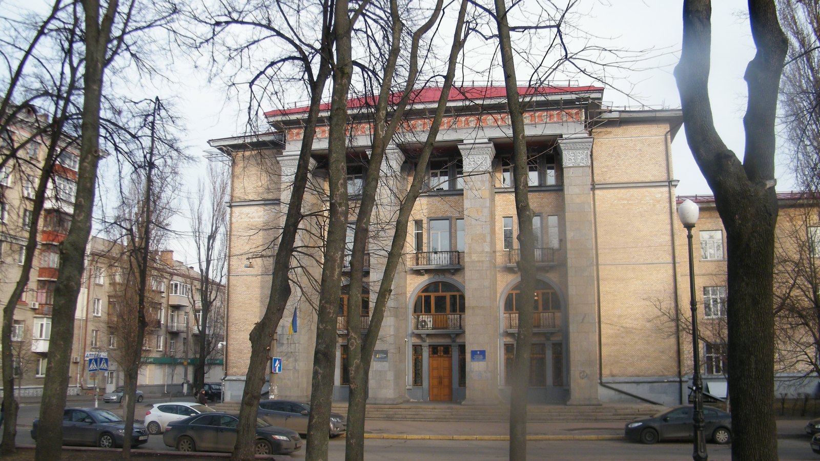 A columned stone office building behind bare trees, with a small Ukrainian flag by the entrance