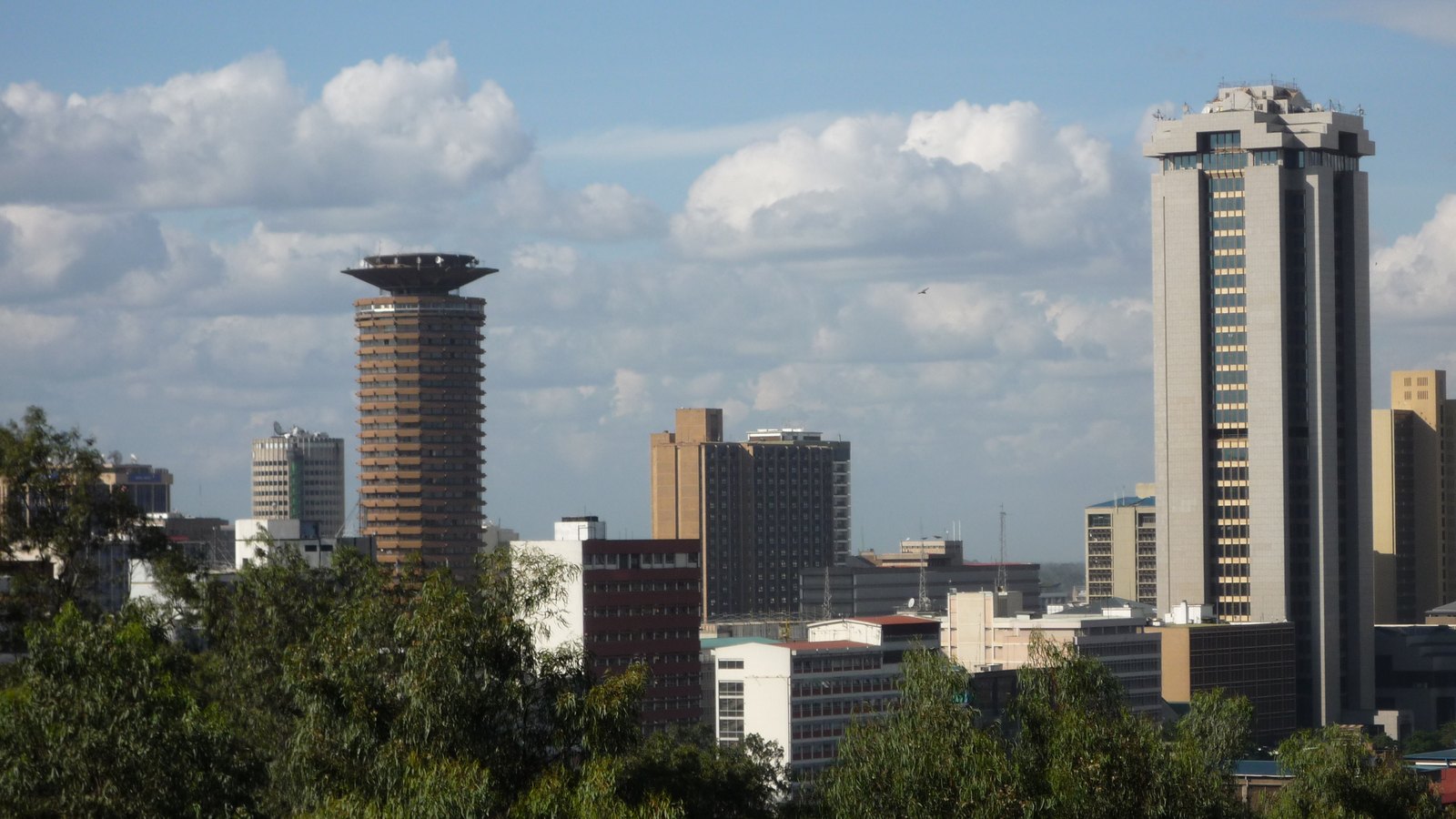 A skyline of office towers, with a tall cylindrical tower, above a band of trees