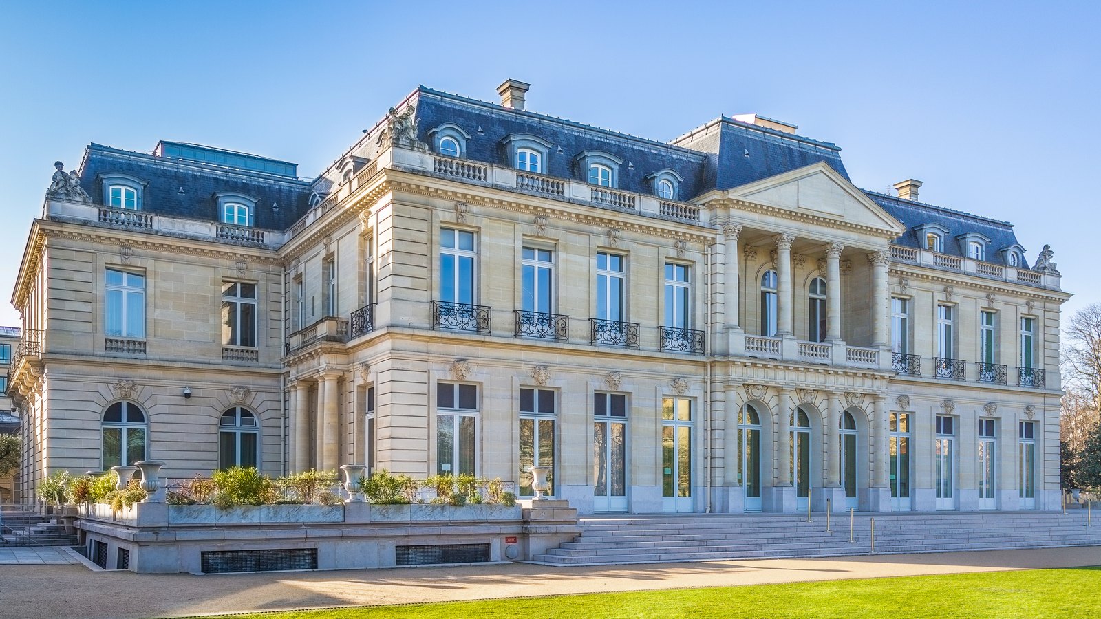 A grand cream stone chateau with a slate mansard roof behind a terrace, under a blue sky
