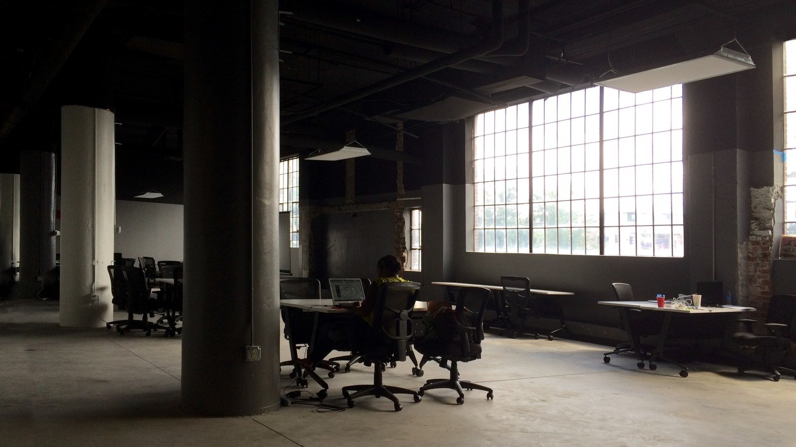 A lone worker at a desk in a dim open-plan office with tall windows and concrete pillars