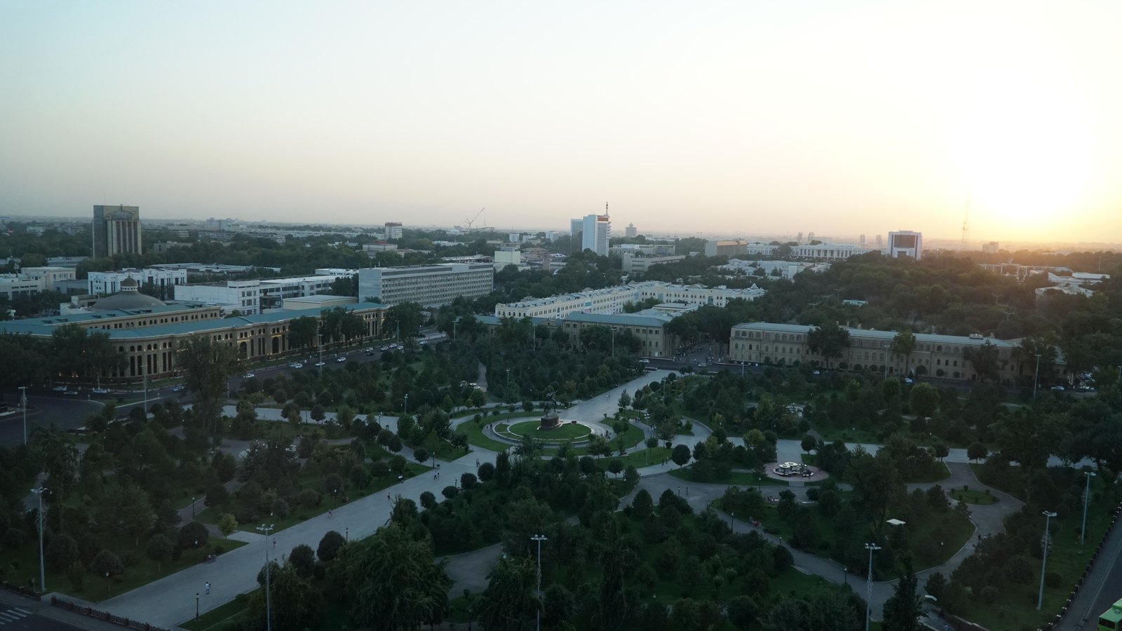 A wide park with tree-lined paths surrounded by large buildings, seen from above in the evening light