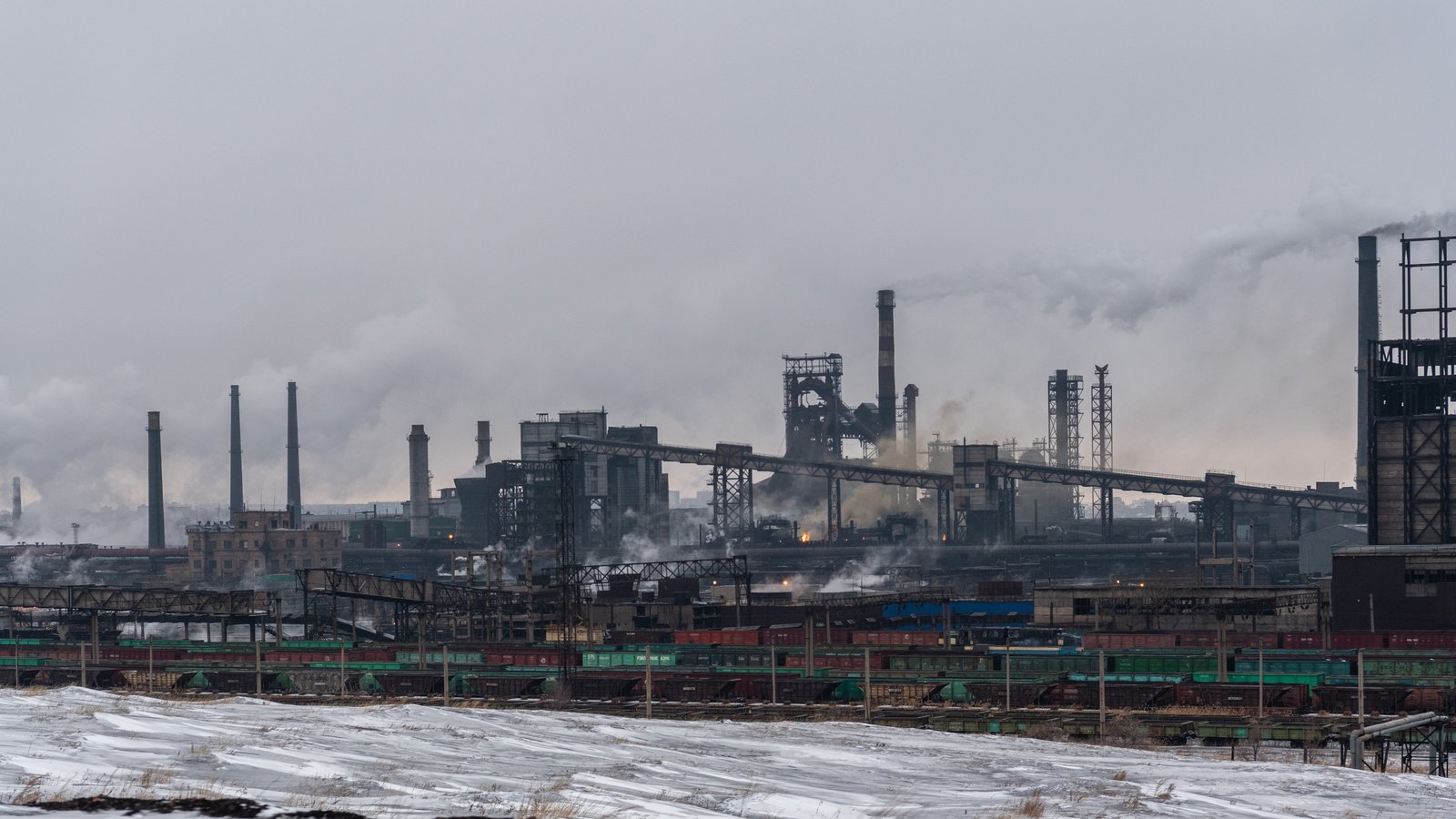 A steelworks with blast furnaces and chimneys under a grey winter sky, beyond snow-covered ground