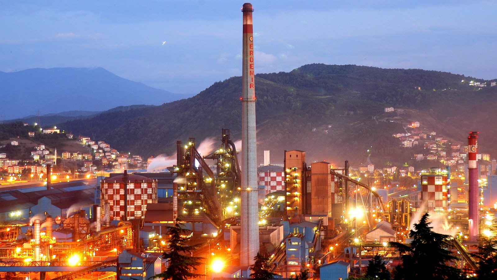 A steelworks lit up at dusk, with a tall red and white chimney and wooded hills behind