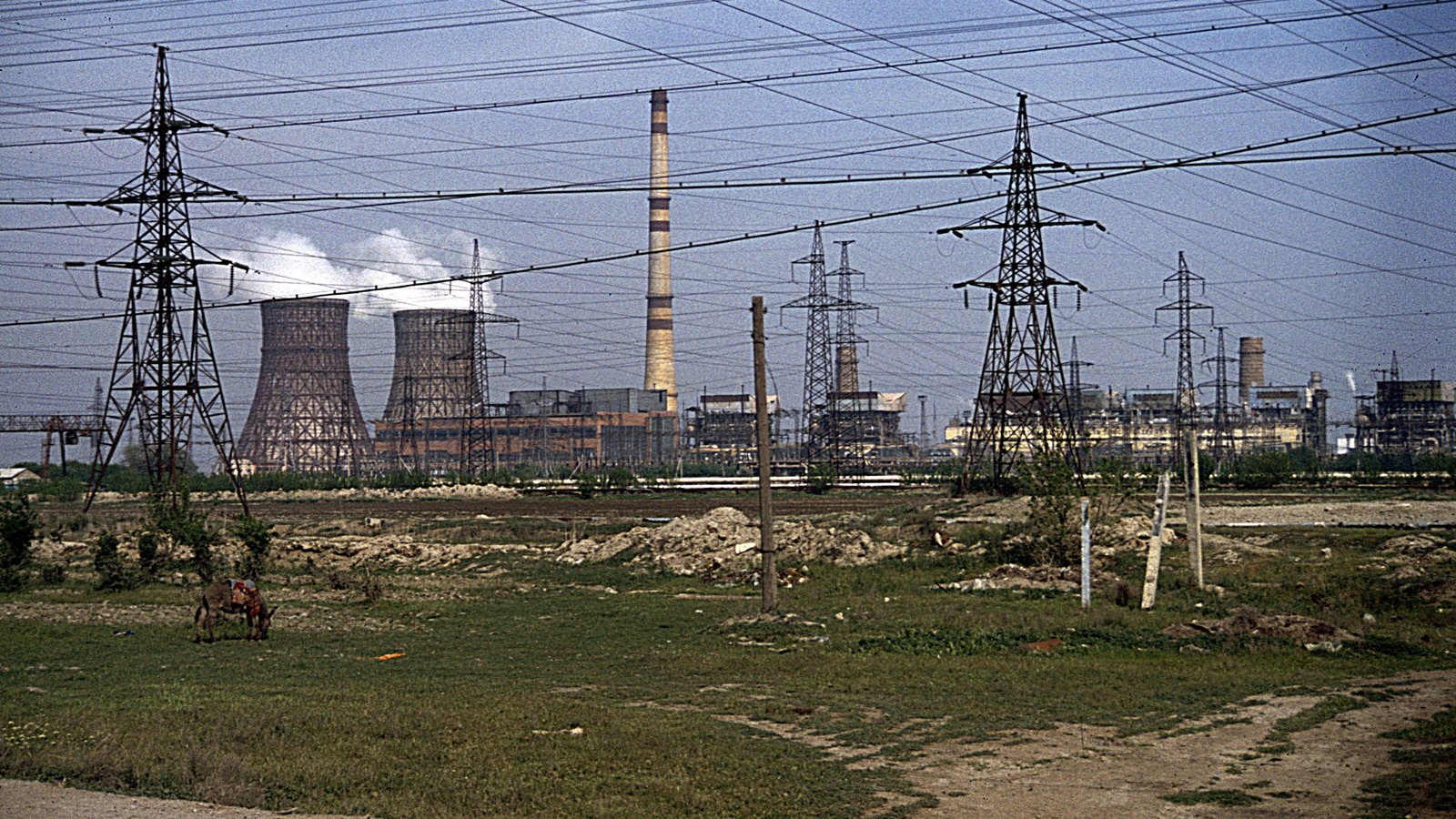 Cooling towers and a tall chimney of a power plant behind high-voltage pylons and open ground