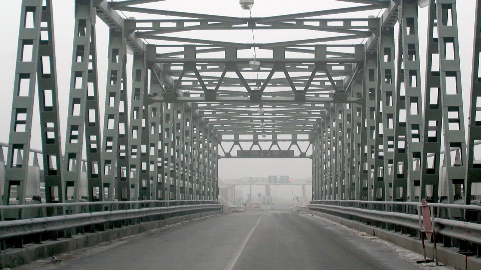 Looking along the road deck of a steel truss bridge towards a border checkpoint