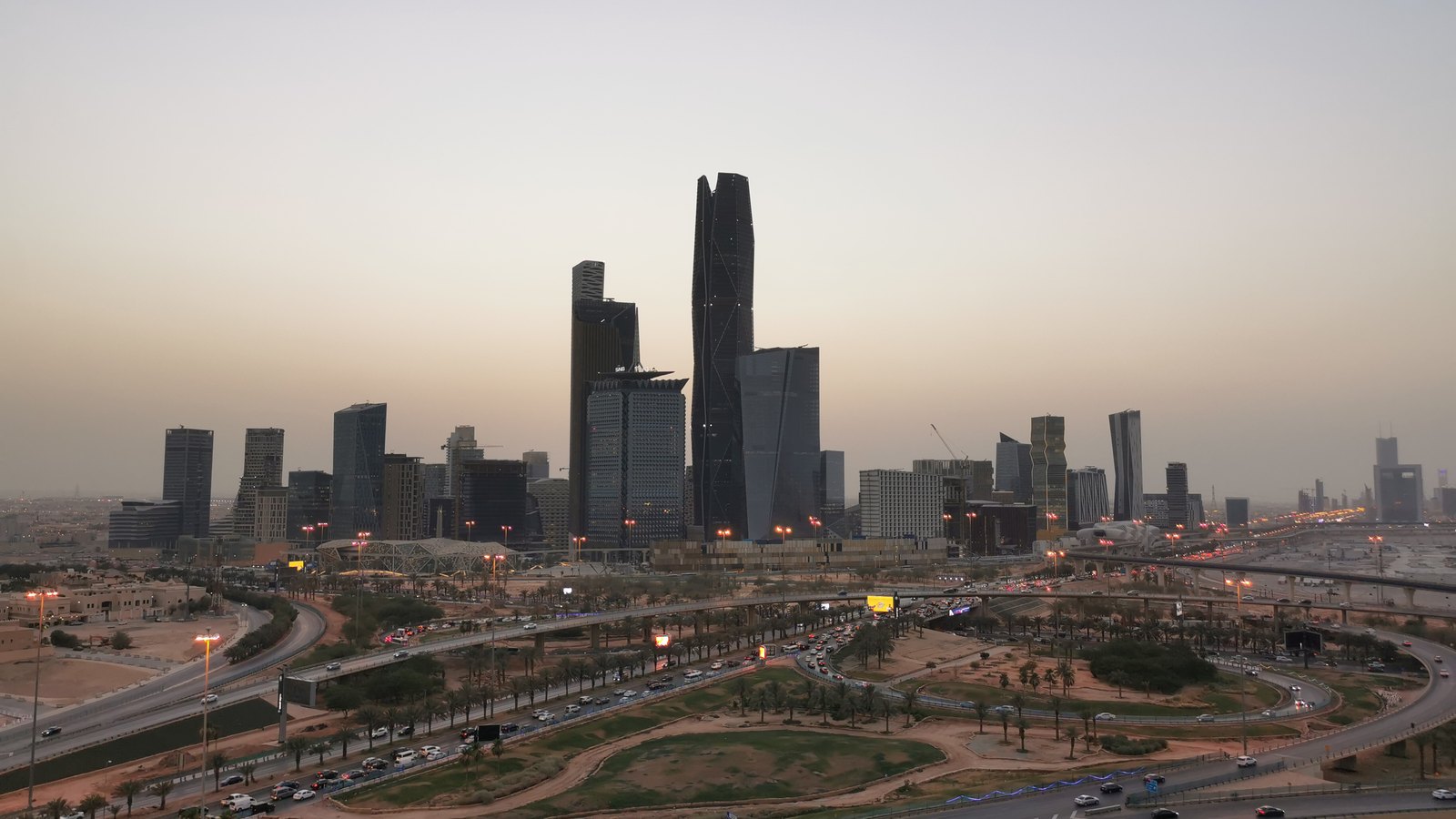 Riyadh financial district towers silhouetted against a pale dusk sky above busy roads, lights and palms