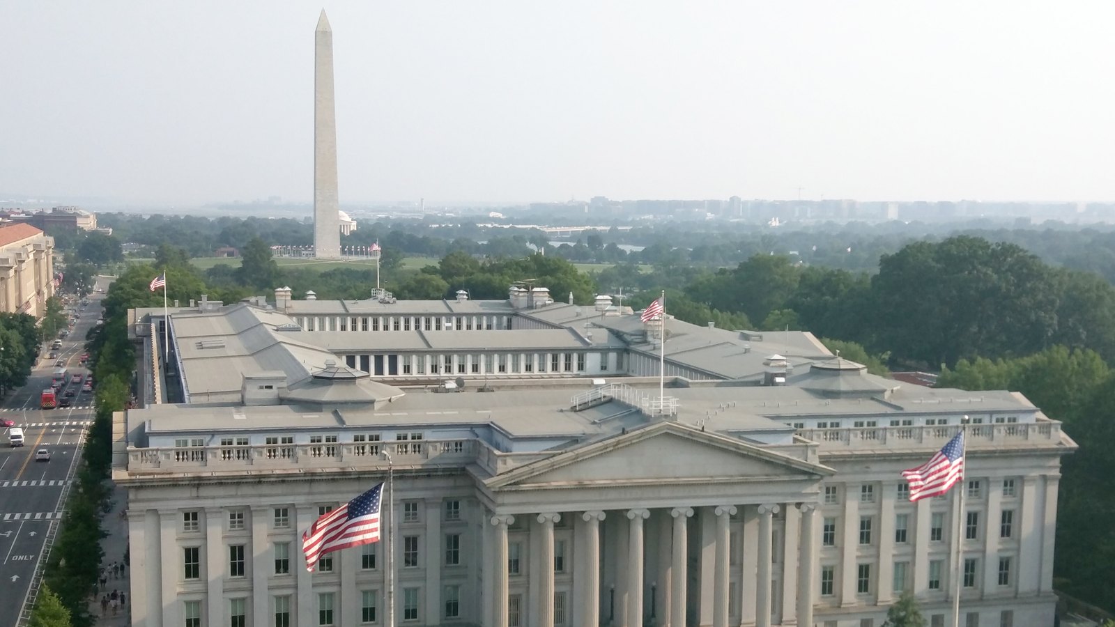 Aerial view of the Treasury Building, its columned front flanked by US flags, with the Washington Monument behind